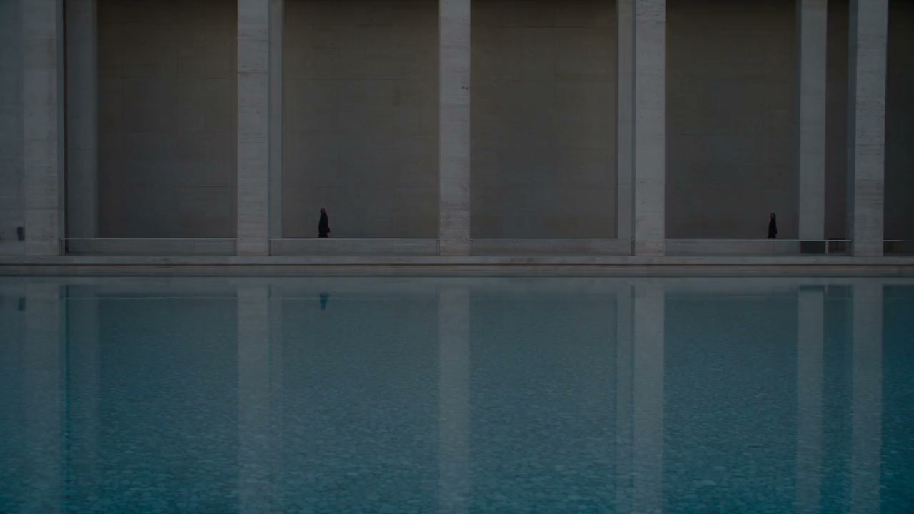 A colonnade of pale stone piers along a still reflecting pool at dusk, two distant figures walking behind the columns.