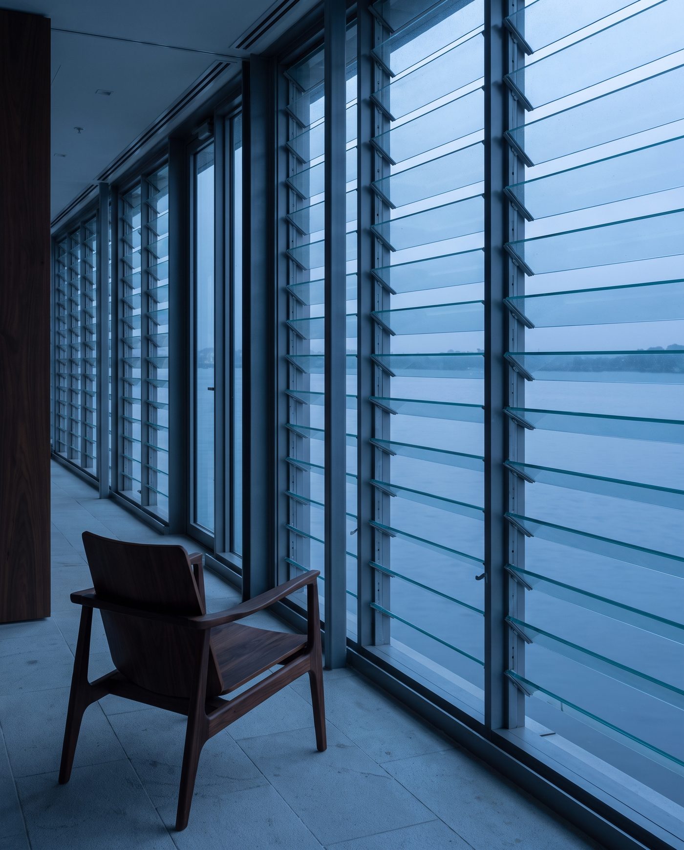 A wall of glass louvres in a quiet office corridor, a single chair turned toward the window.