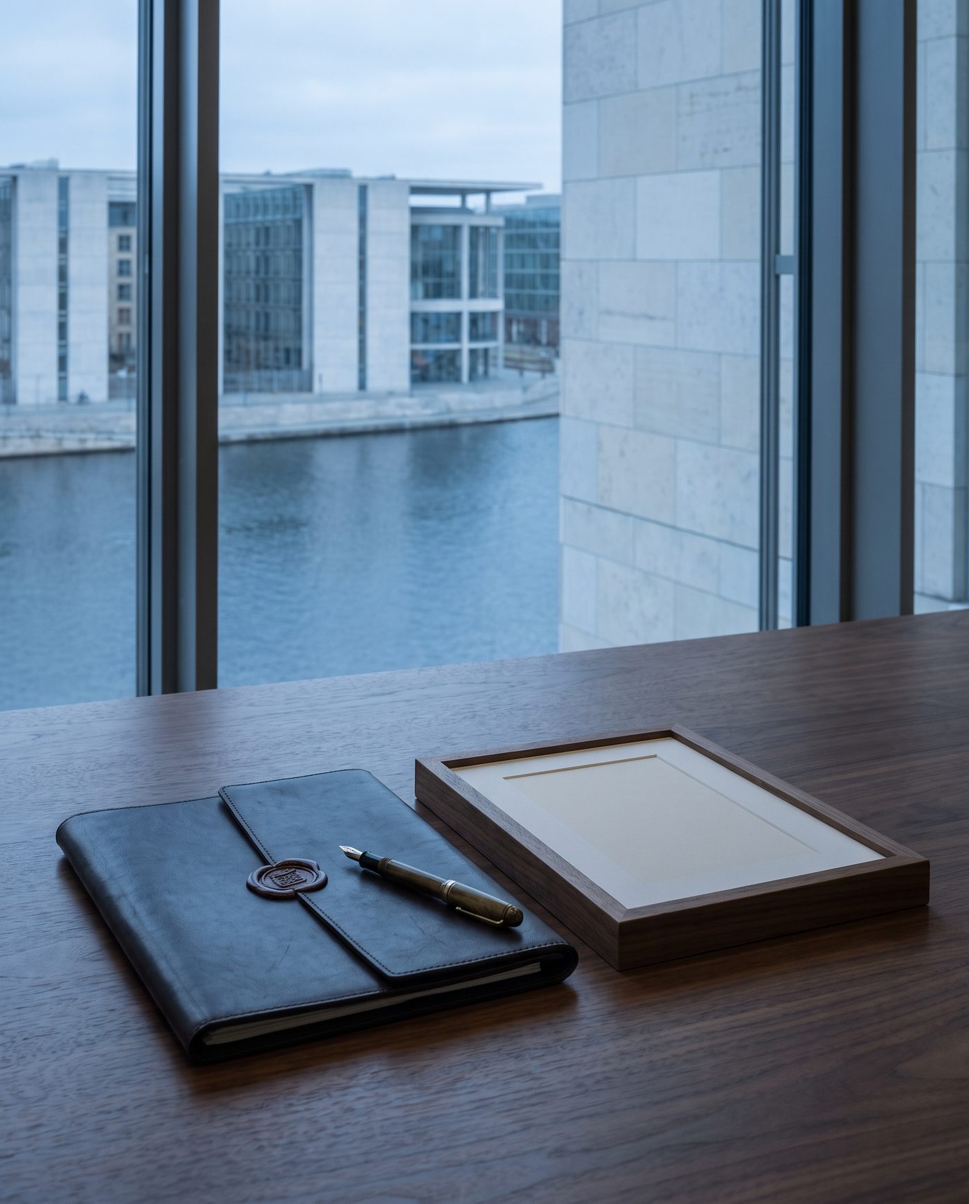 A sealed document folio and a fountain pen resting on a walnut table beside a framed blank photograph.