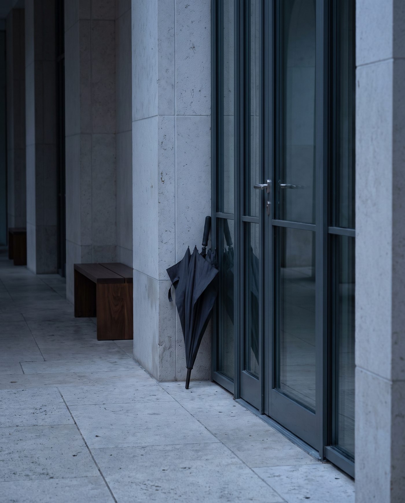 A closed umbrella standing beside a glass door in a quiet stone-floored corridor.