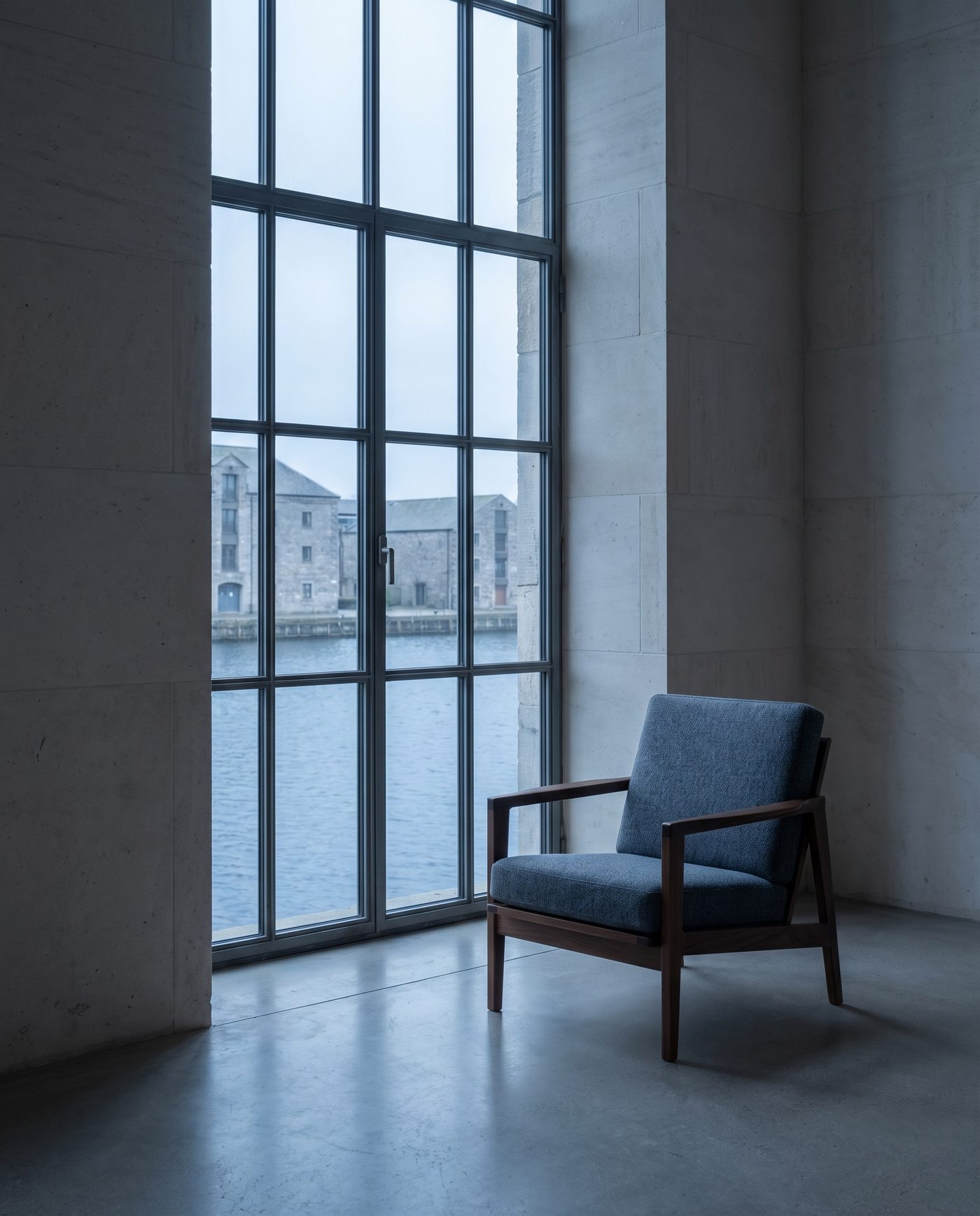 An empty armchair beside a tall window in a quiet pale room.